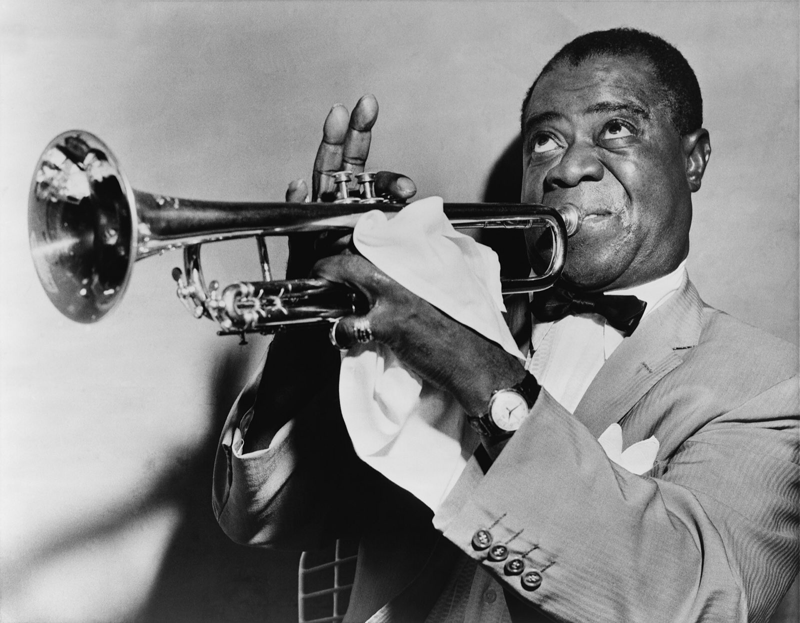Louis Armstrong tocando trompete, de terno claro, com um lenço branco na mão que segura o instrumento.