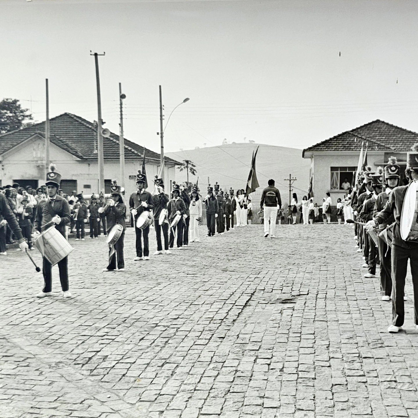 Fanfarra escolar desfilando numa rua de paralelepípedos de cidade pequena, com moradores assistindo da calçada.
