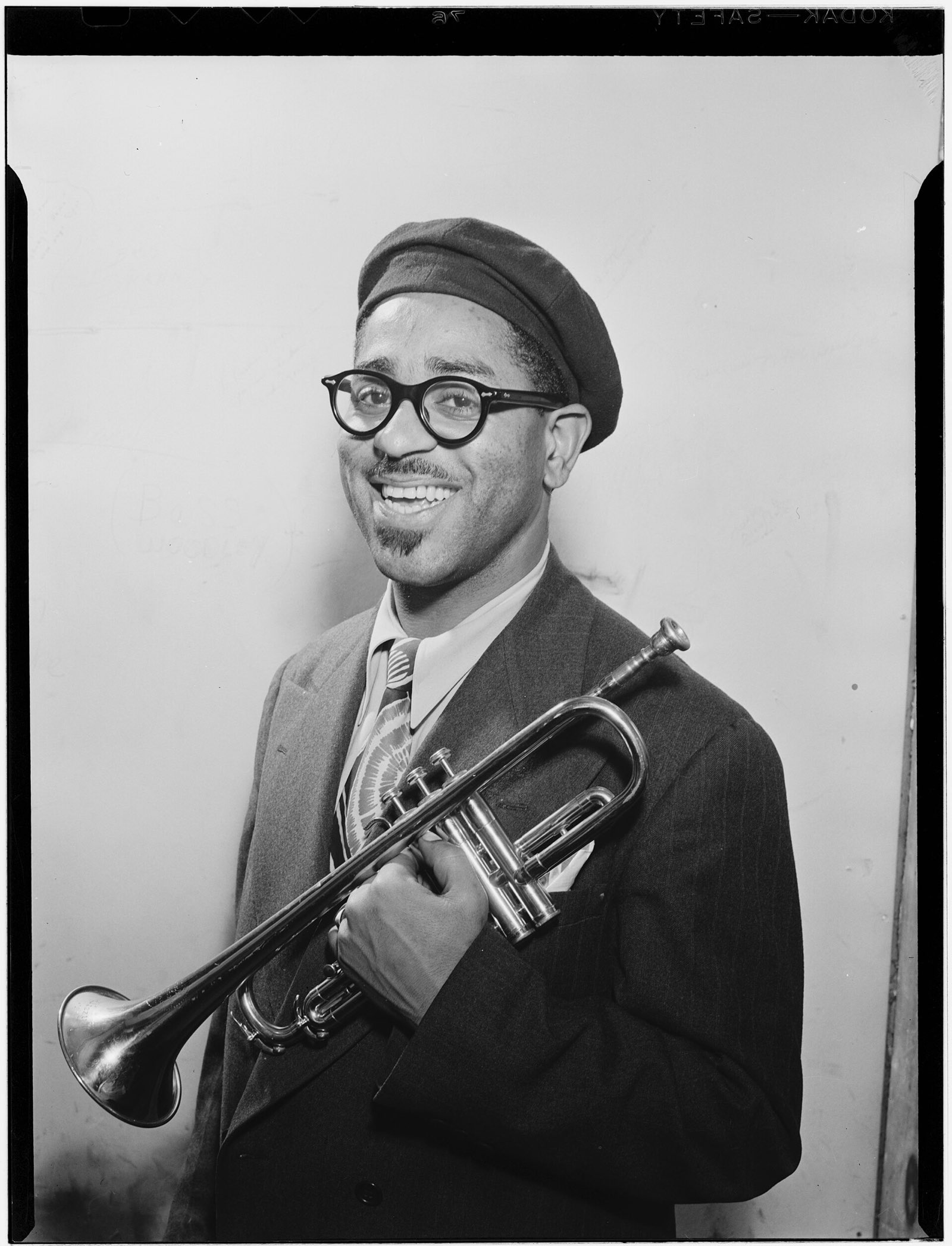 Dizzy Gillespie de boina e óculos redondos, sorrindo, com o trompete apoiado no braço.