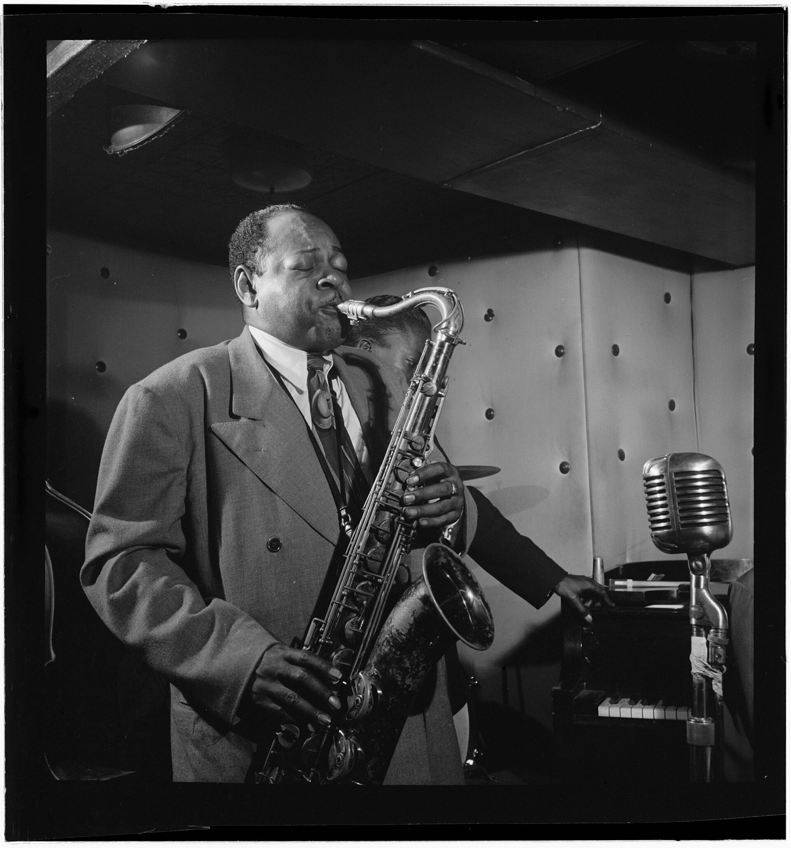 Coleman Hawkins tocando saxofone tenor num clube, de olhos fechados, com um microfone antigo ao lado.