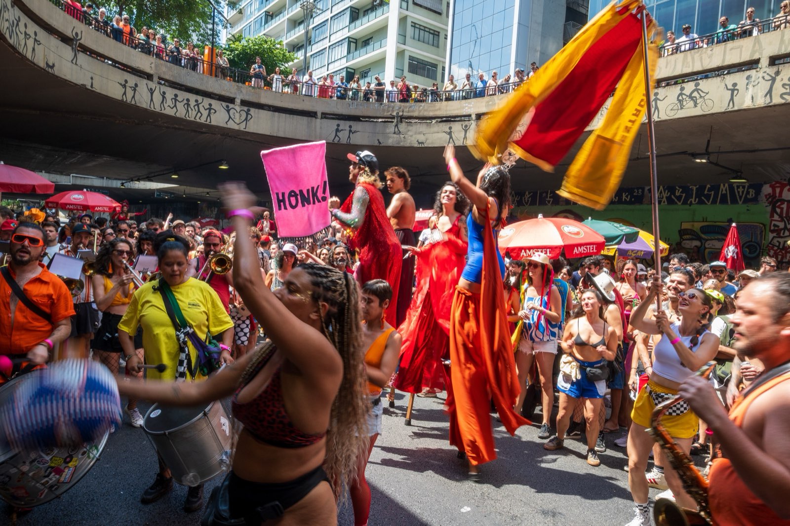 Fanfarra e público lotando um túnel em São Paulo durante o HONK! SP: uma placa escrita HONK!, uma bandeira vermelha e amarela, artistas em pernas de pau e caixas de percussão no meio da multidão.