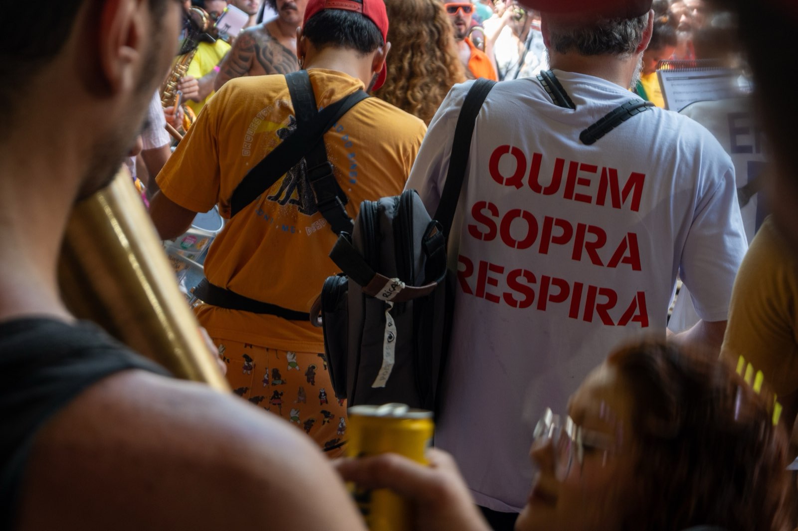 De dentro da fanfarra: o latão de um sopro carregado no ombro em primeiro plano e, à frente, as costas de um músico com uma camiseta onde se lê “quem sopra respira”.