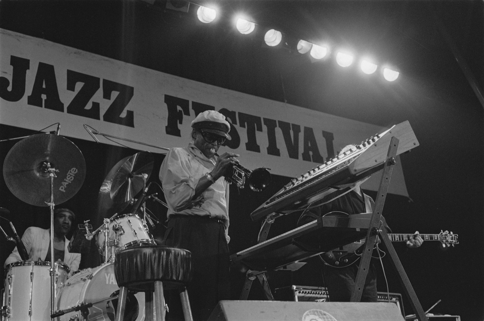 Miles Davis tocando trompete num palco de festival, de boné e óculos escuros, cercado de teclados e bateria.